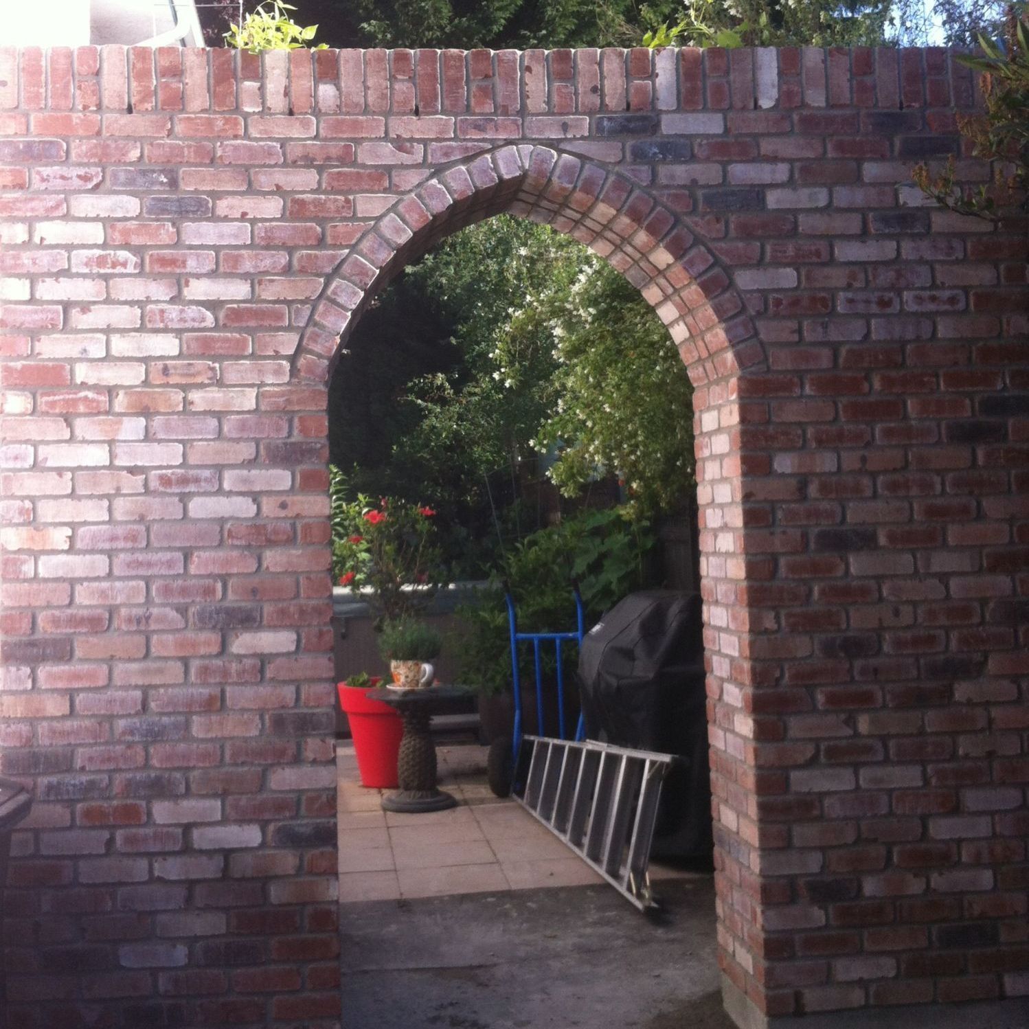 Brick wall with arched doorway leading to a patio with a red pot and a ladder.