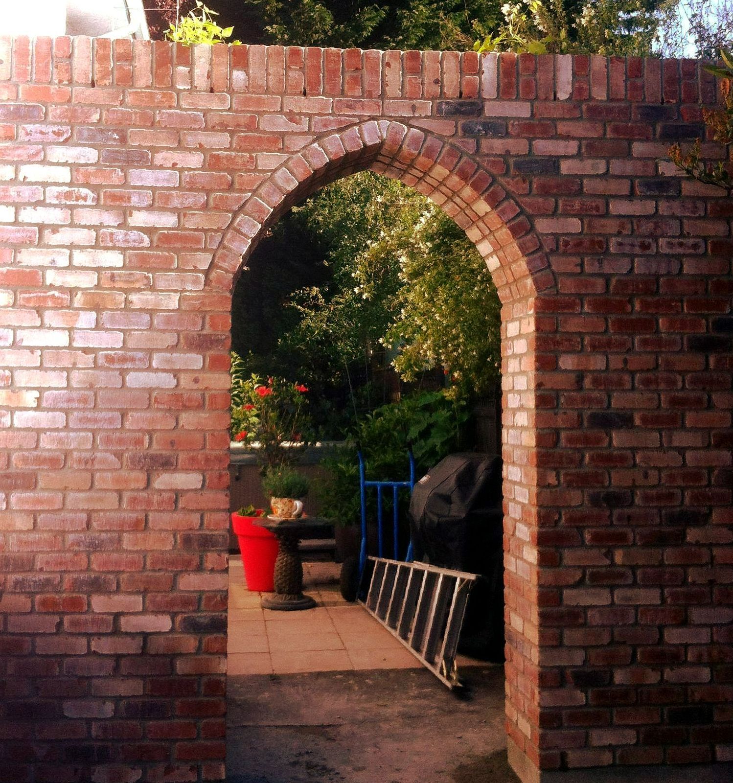 Brick archway in a wall, framing a view of a patio with a table, plants, and a ladder.