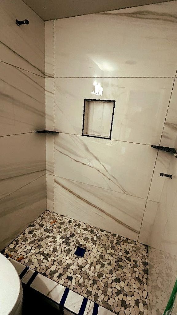 Shower with marble-like tile walls and a pebble floor, a built-in shelf and square niche.