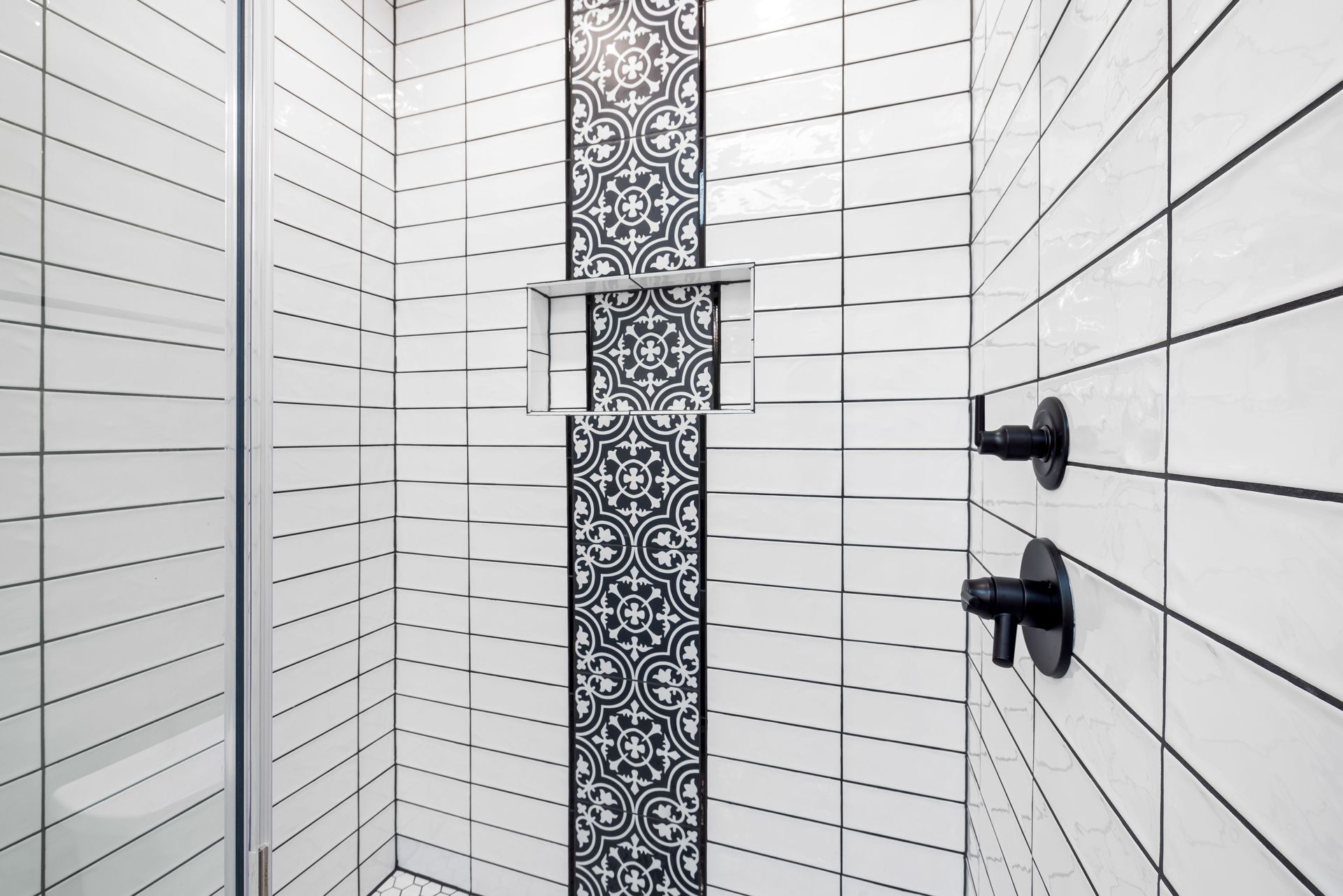 Narrow white tiled shower stall with black shower floor and small corner shelves
