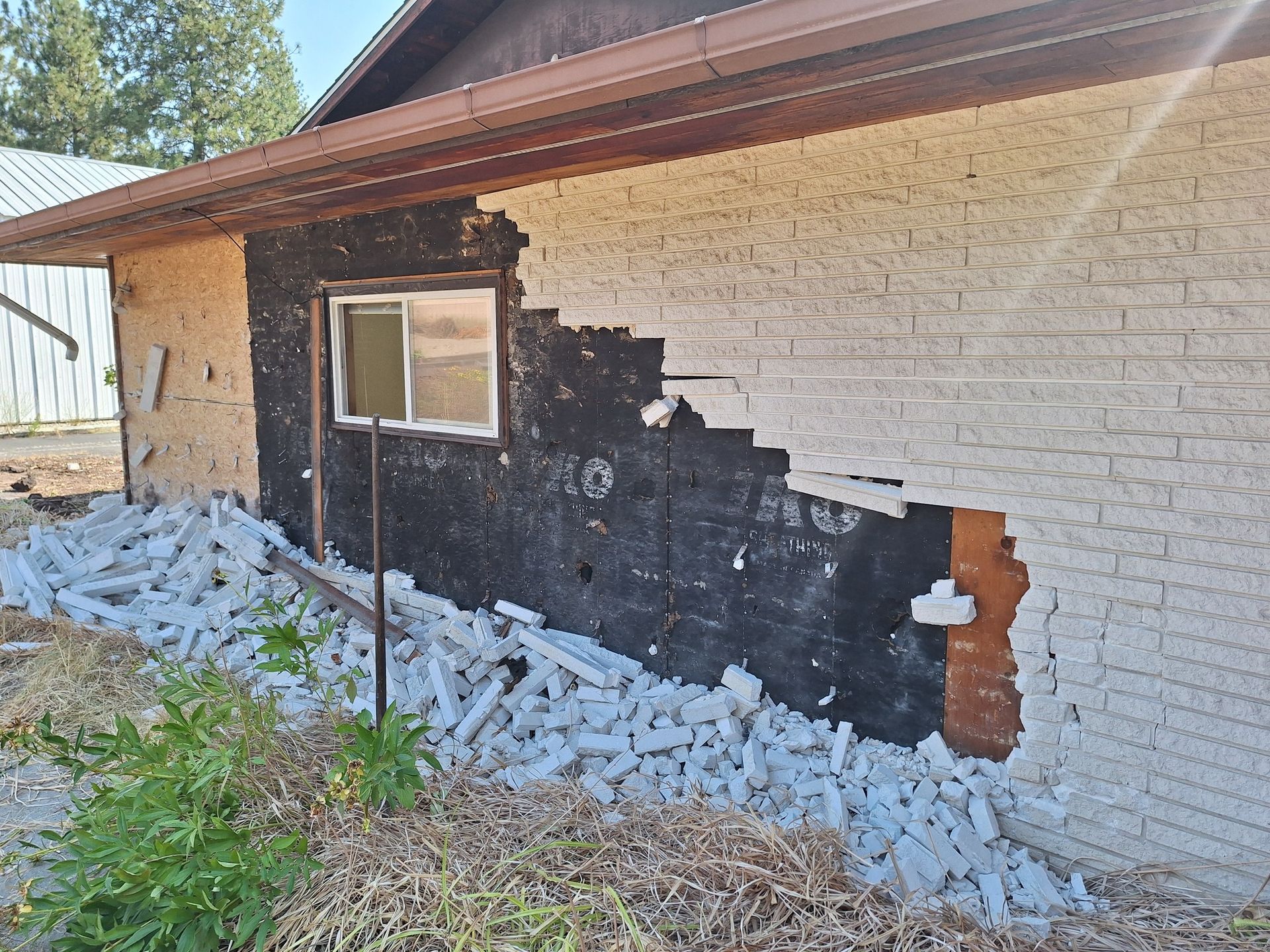 Exterior wall of a house with collapsed light-colored brick veneer, exposing the black building paper underneath.