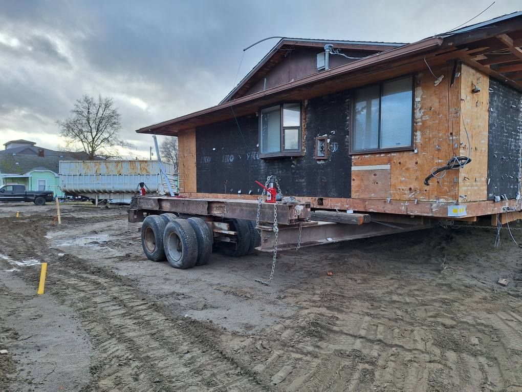 A wooden house structure is elevated on steel beams and a multi-axle trailer, staged on a muddy construction lot.