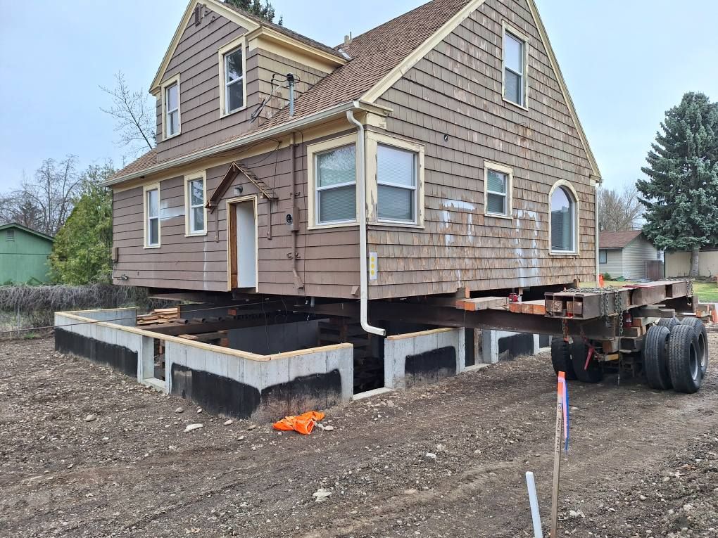 A tan two-story house is being moved onto a new concrete block foundation, supported by steel beams and a trailer.