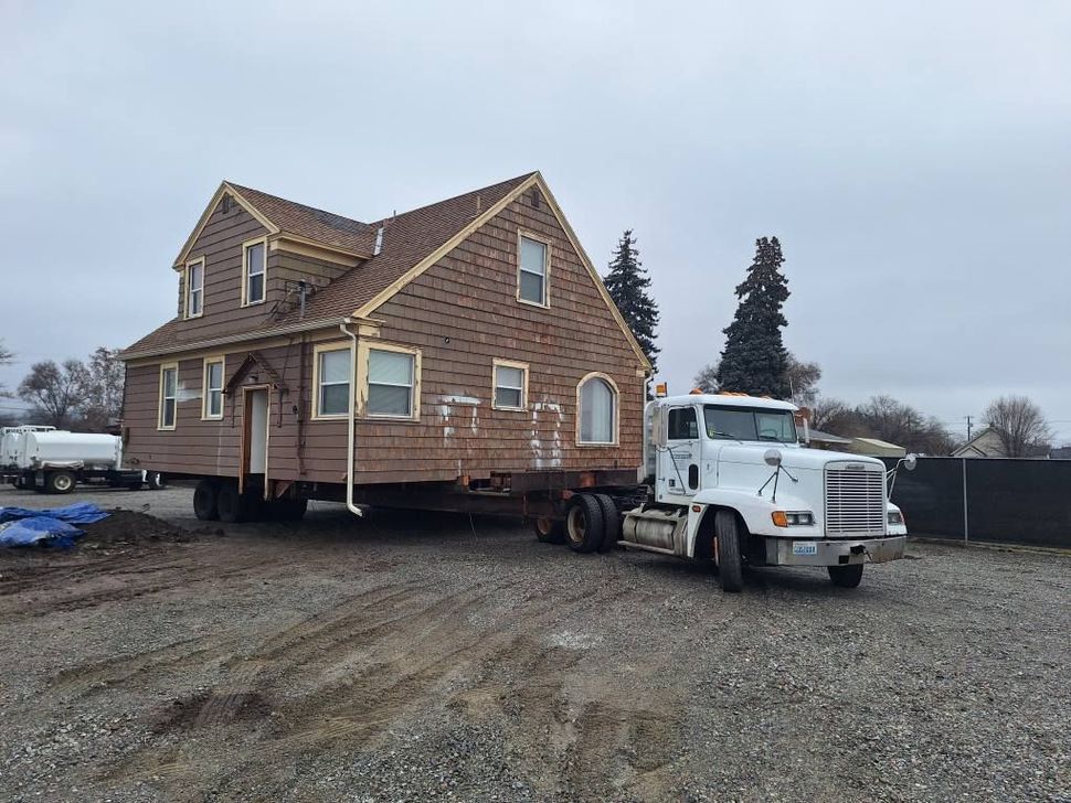 A white semi-truck hauls a two-story tan house, stripped of its siding, across a gravel lot on a cloudy day.