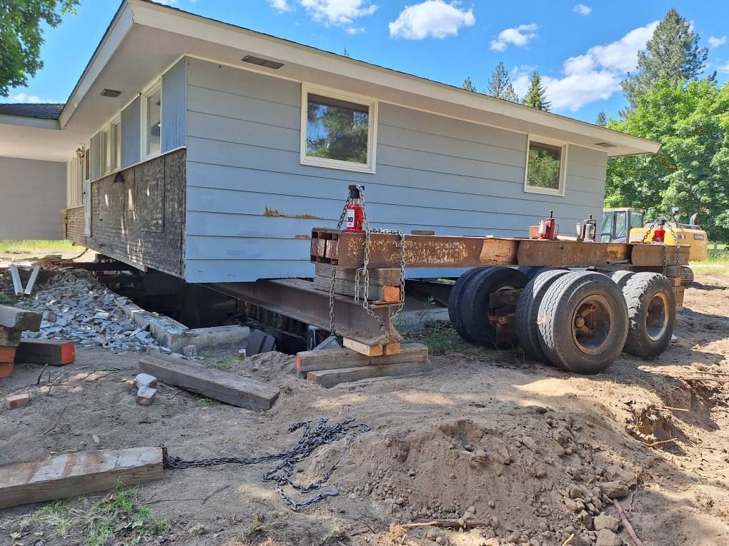 A blue house raised on steel beams and a multi-axle trailer for transport on a dirt lot.