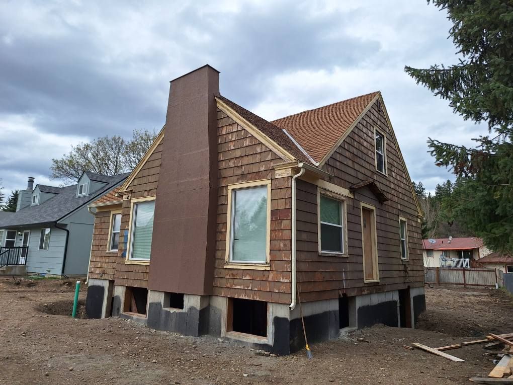 A house under renovation with exposed wood siding, a prominent brown chimney, and a bare foundation in a dirt yard.