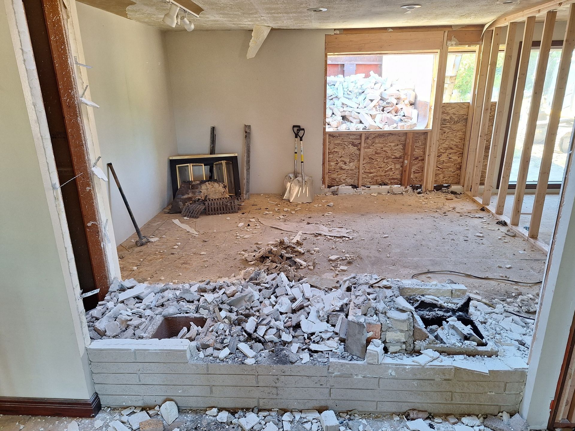 An interior room under construction with debris on the floor, a partially demolished brick wall, and exposed wall studs.