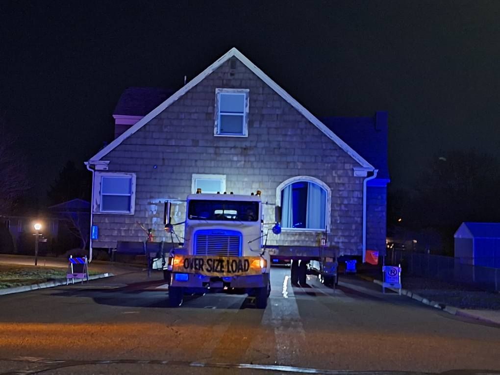 A white semi-truck pulls a large, single-story stone house down a dark residential street at night.