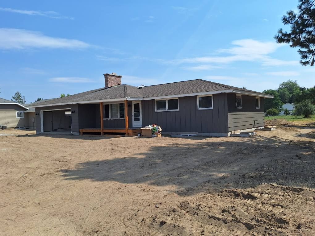A one-story dark gray house with a new wooden porch and roof, set against a backdrop of bare dirt and blue sky.