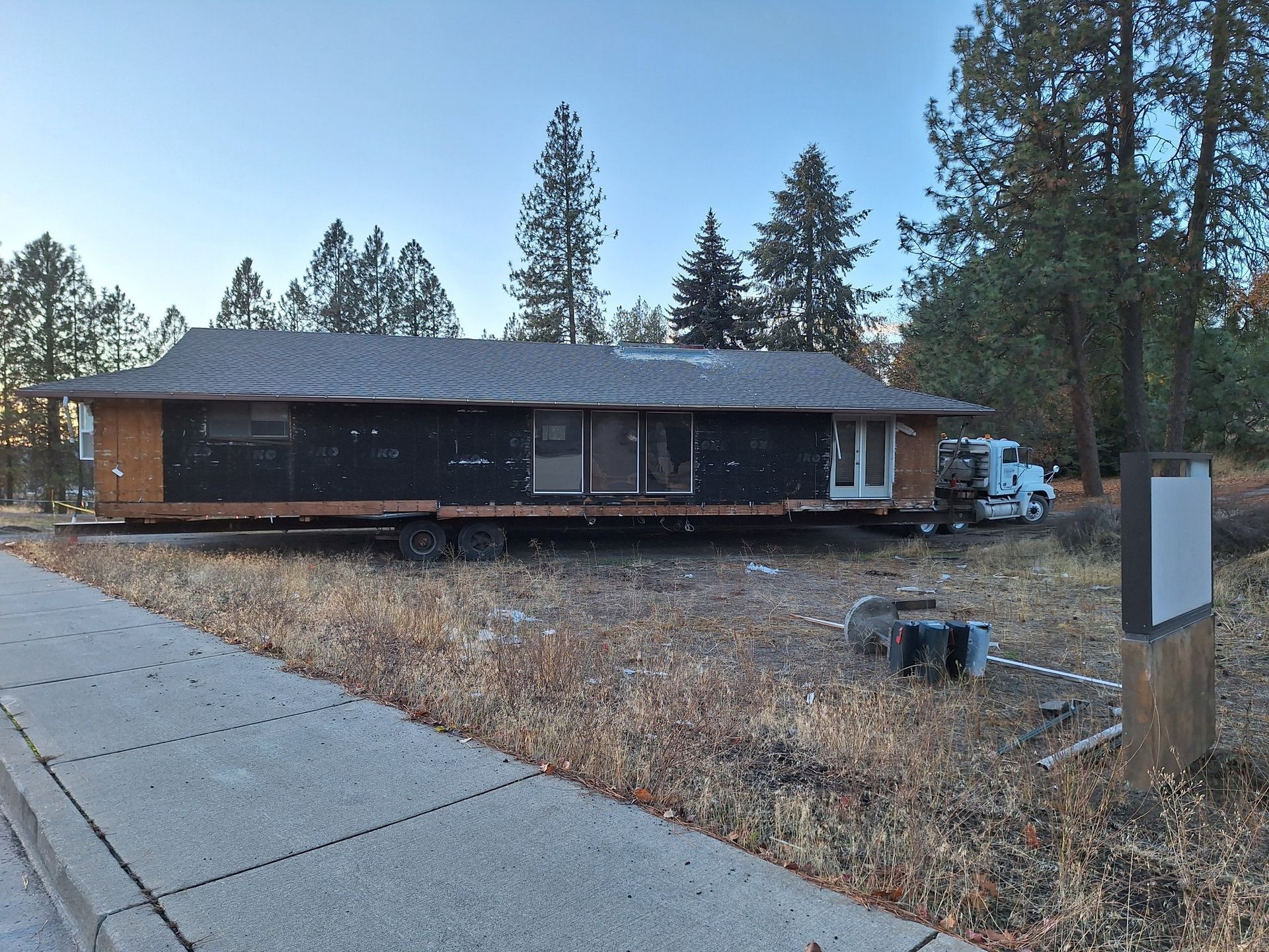 A mobile home sits on a trailer in a rural, wooded area next to a paved path, with a white truck parked nearby.