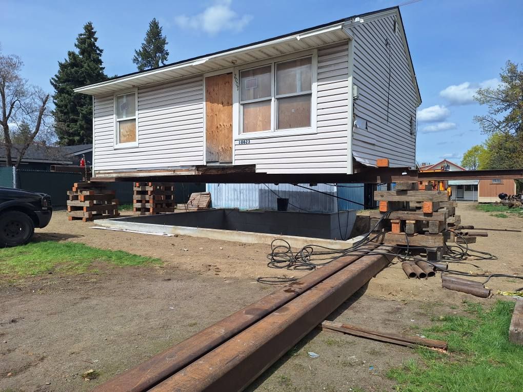 A white house lifted on steel beams and wooden cribbing blocks, staged for relocation or foundation work.