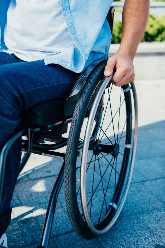 Persona in sedia a rotelle all'aperto, con la mano sul volante, che indossa una camicia azzurra e jeans.