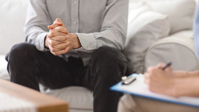 Durante una seduta di consulenza, due persone siedono su un divano bianco: una con le mani giunte e l'altra che prende appunti.