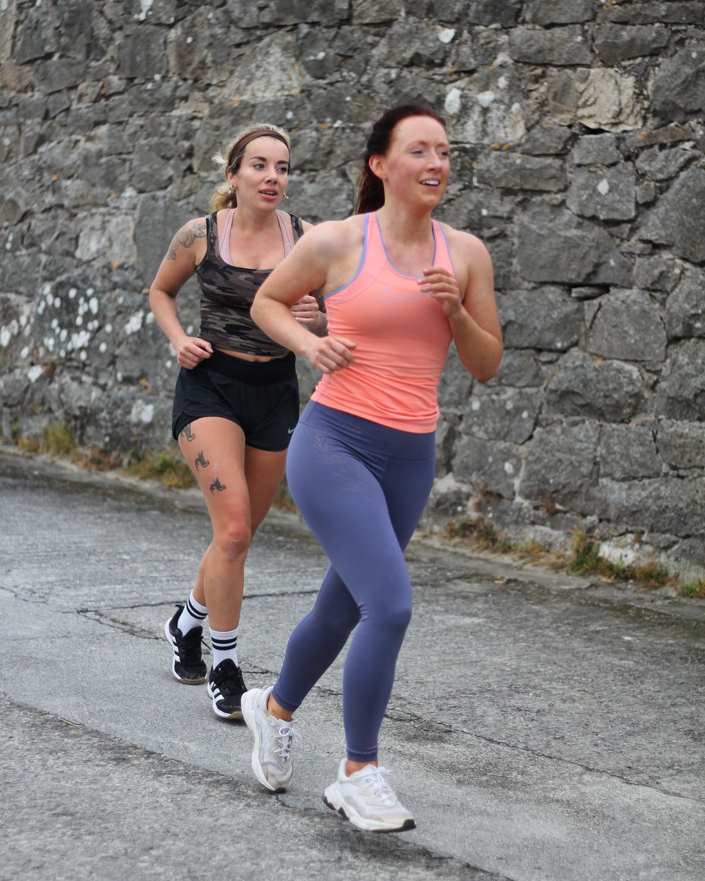 Two women are running down a street in front of a stone wall.