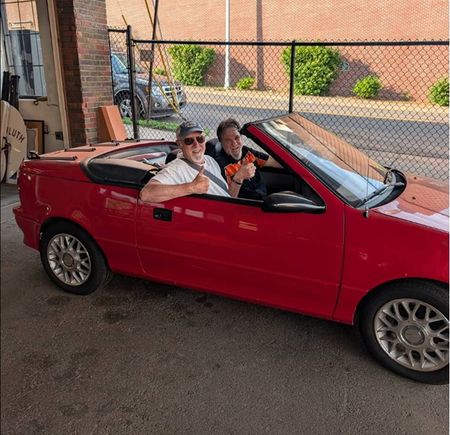 Two people sitting in a red convertible car in a garage, both smiling and waving. | Exodus Motor Works LLC