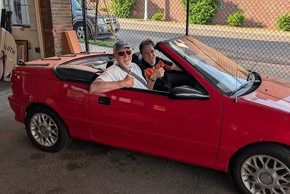 Two people riding in a red convertible parked by a chain-link fence | Exodus Motor Works LLC