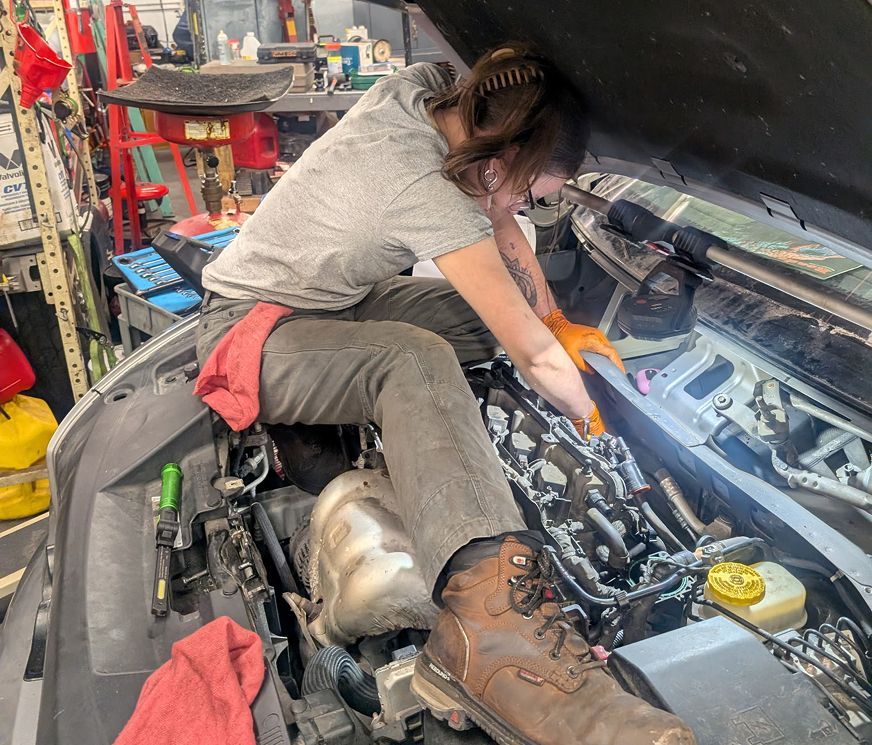 Mechanic working under a car hood in a garage, leaning over the engine with tools and red shop rags nearby | Exodus Motor Works LLC
