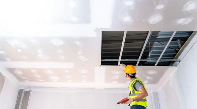 Worker in yellow hard hat inspecting a ceiling with exposed tiles and bright spotlights in a room under renovation