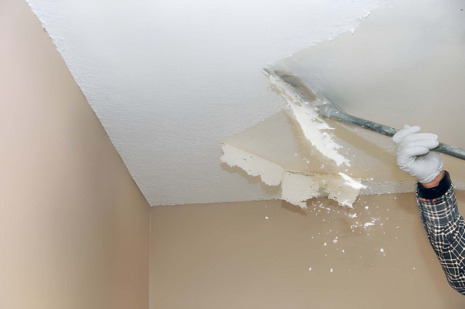 Person scraping damaged paint from a white ceiling with a putty knife in a beige room