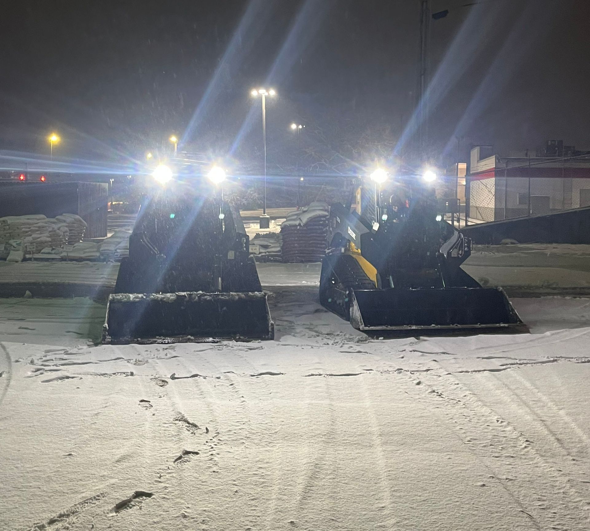 Two skid steer loaders with bright headlights clearing snow at night.