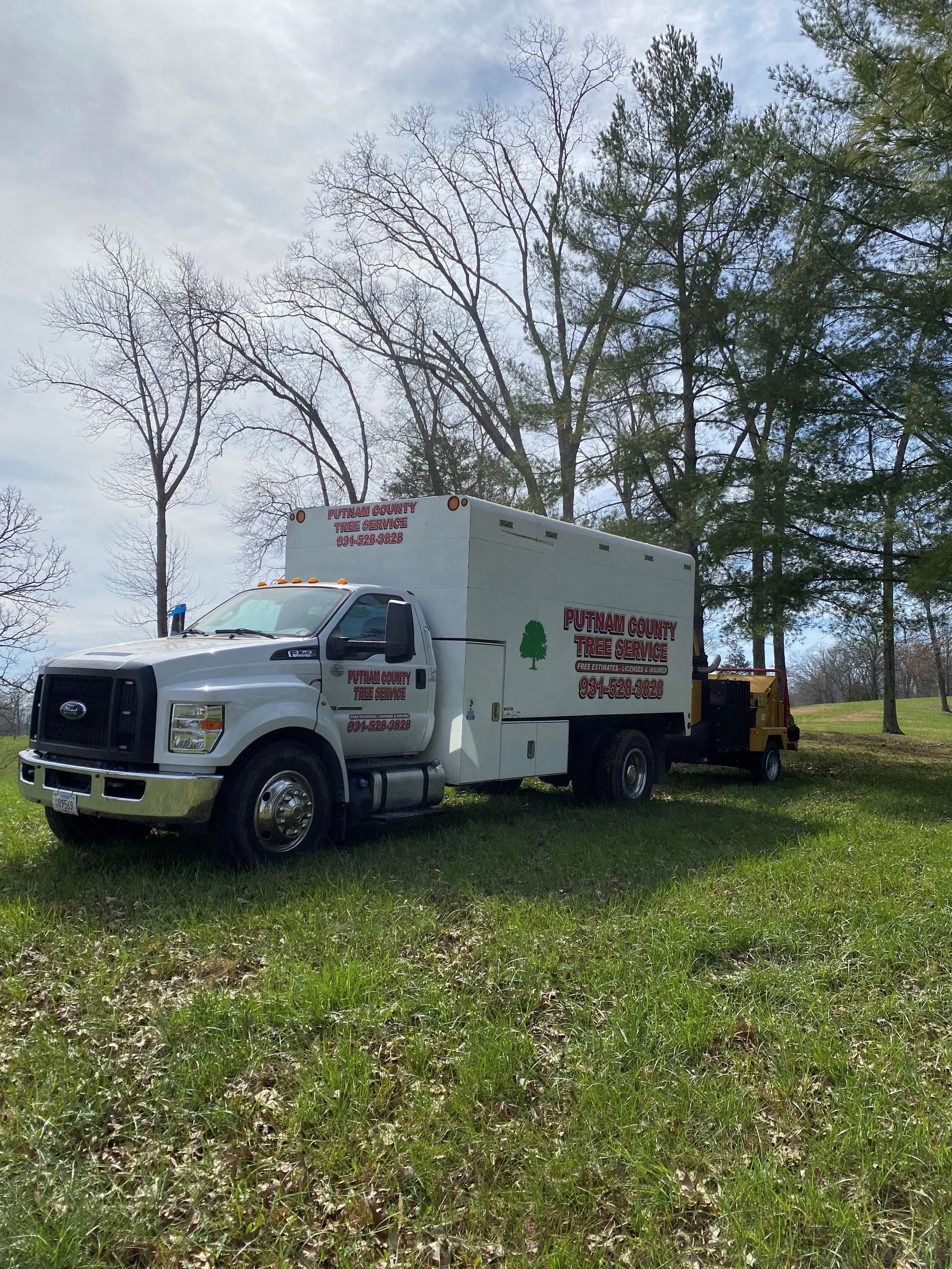 White tree service truck and stump grinder on a grassy field with trees under a cloudy sky.
