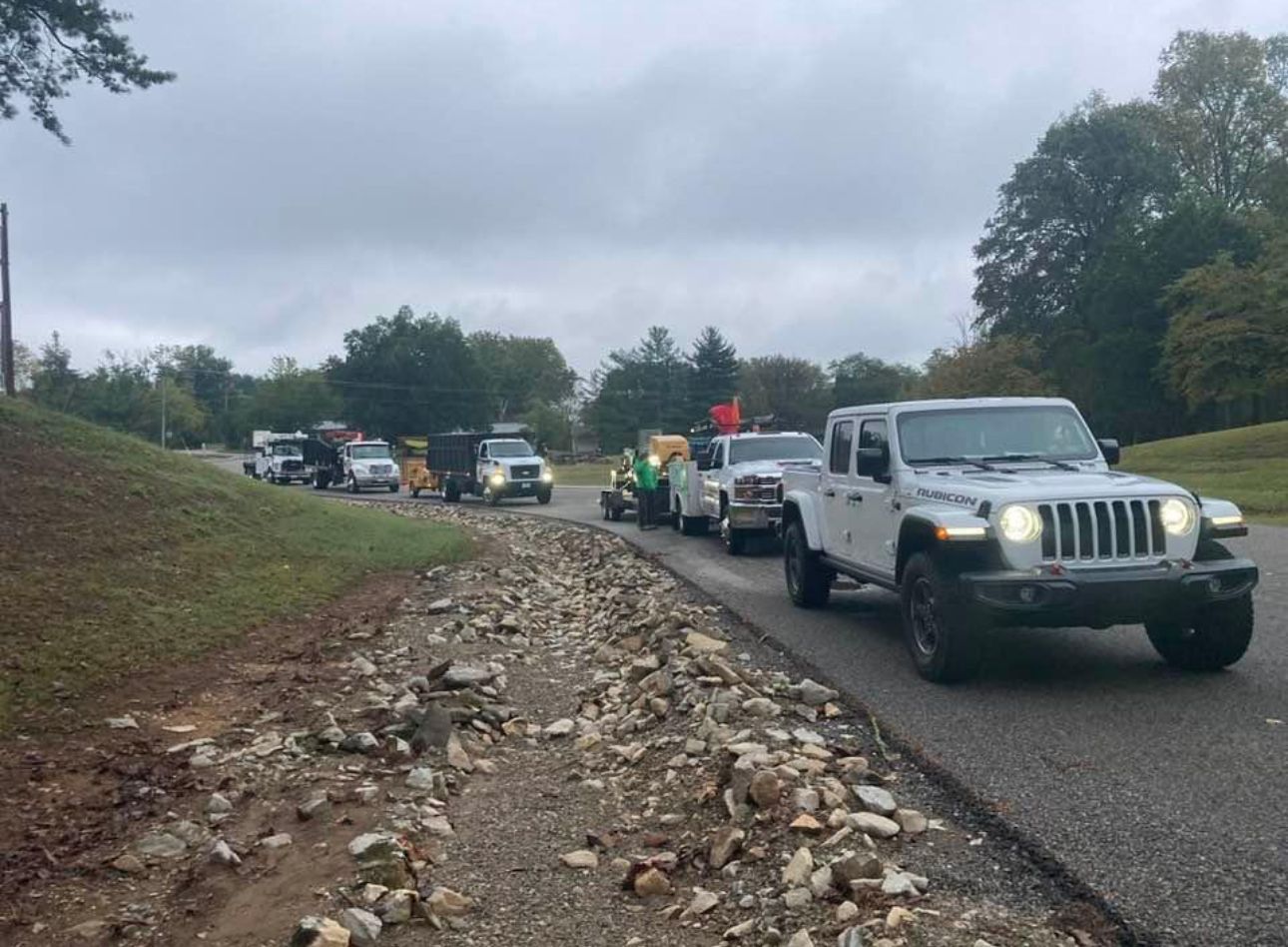Line of off-road vehicles on a paved road next to a rocky area under an overcast sky.
