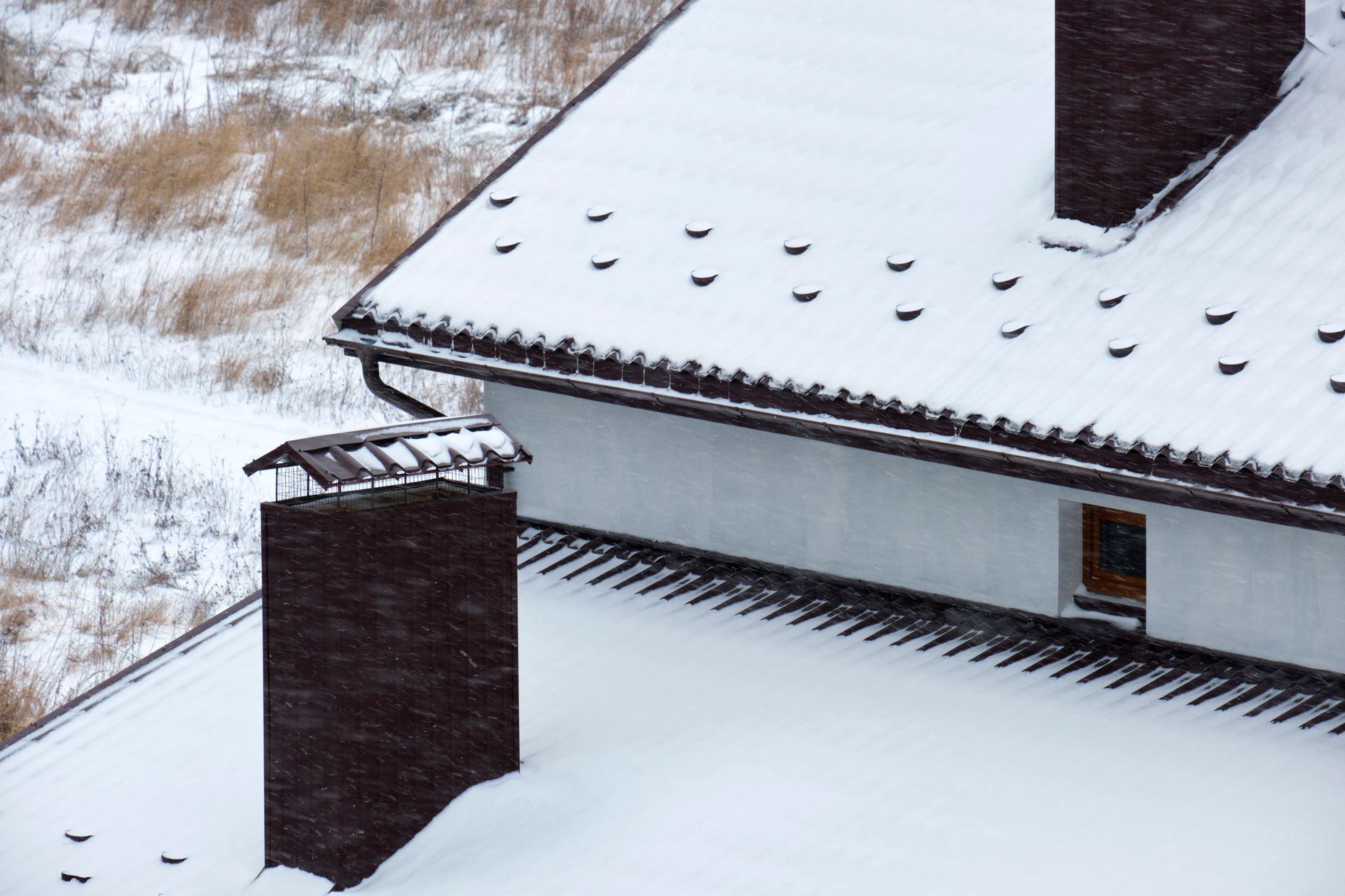 Snow-covered house roof with a brick chimney and a small window.