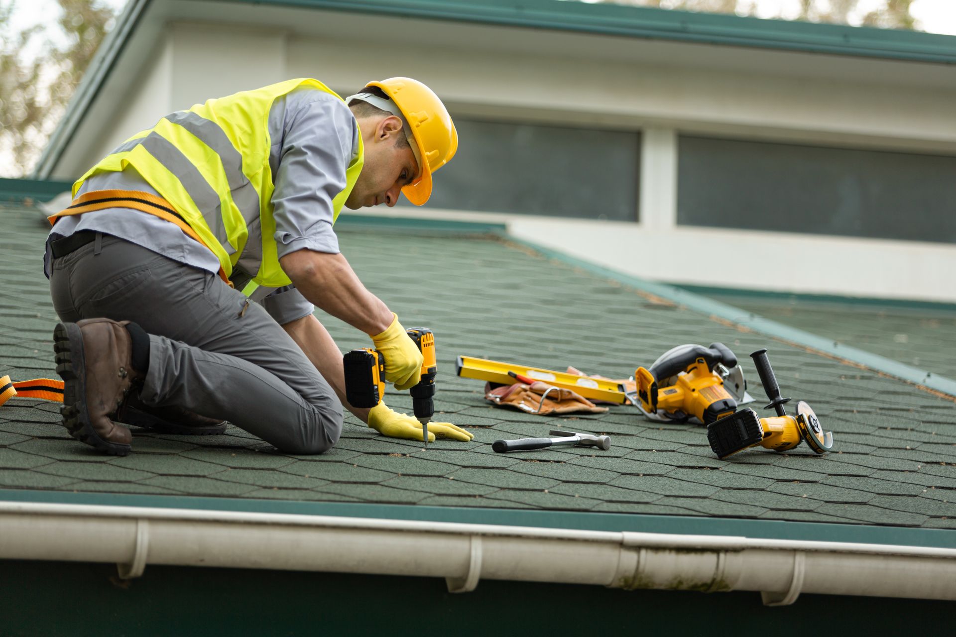 Worker in yellow safety vest and hard hat kneels on a roof, using tools to repair green shingles.