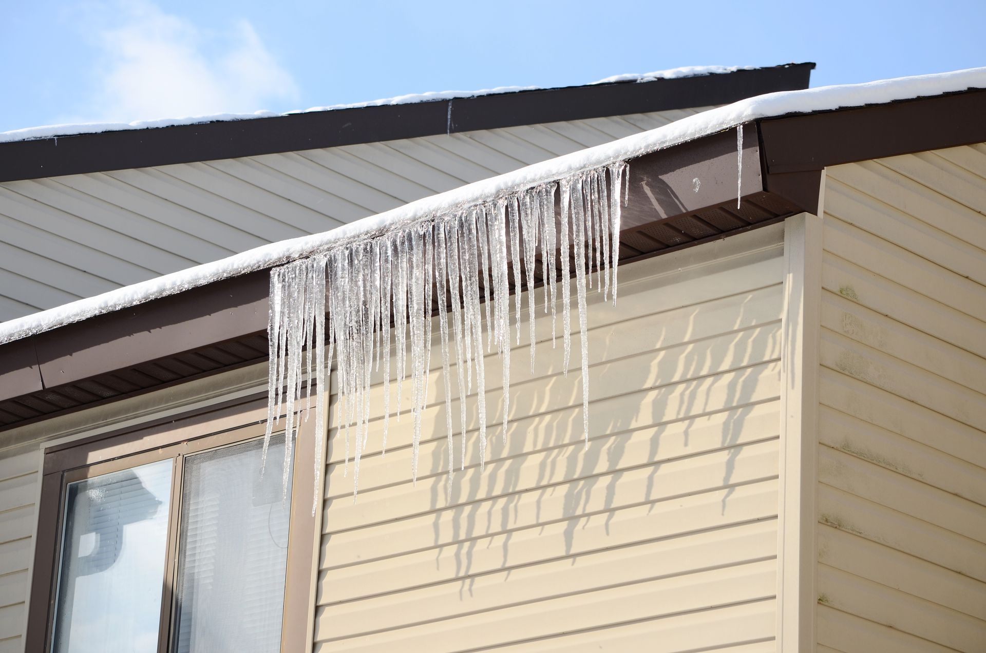 Icicles hanging from a brown roof overhang on a light-colored building, against a blue sky.