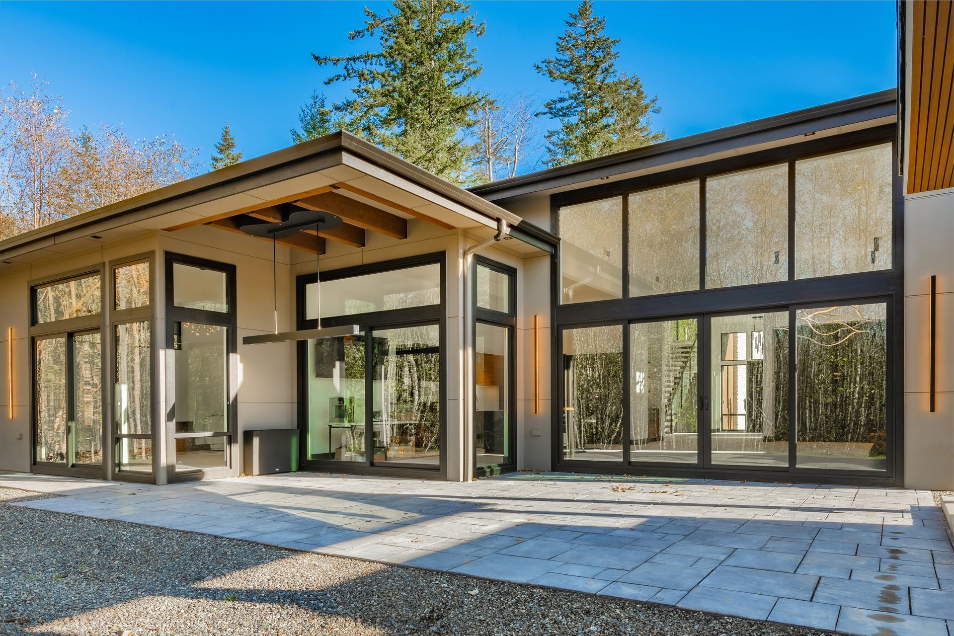 Modern house with large windows, light exterior, and stone patio. Trees in the background.