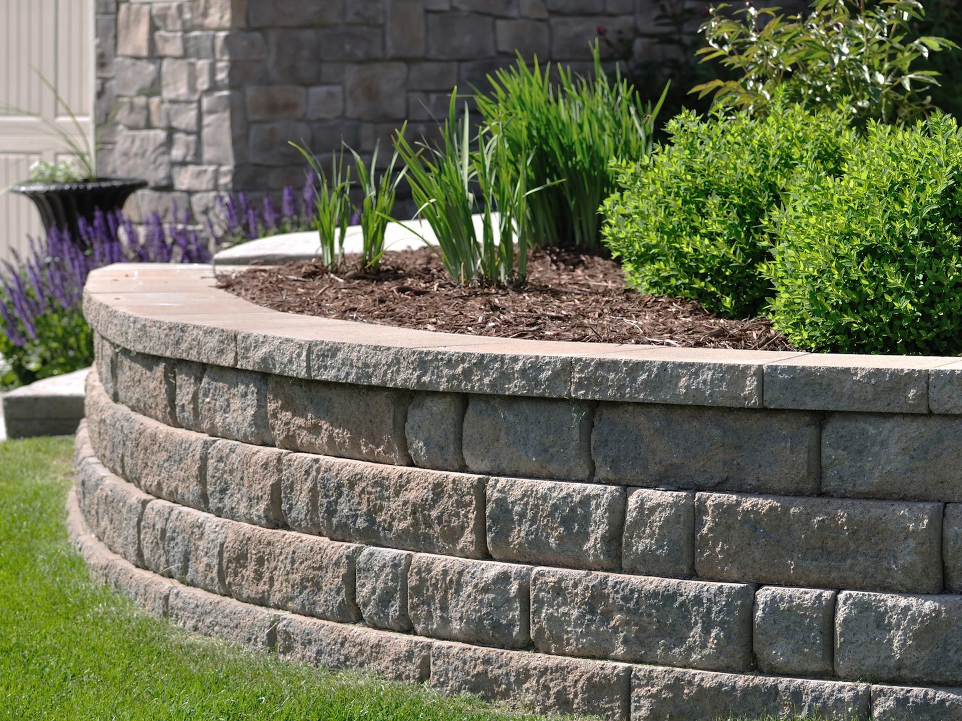Curved gray stone retaining wall with green plants and grass lawn.