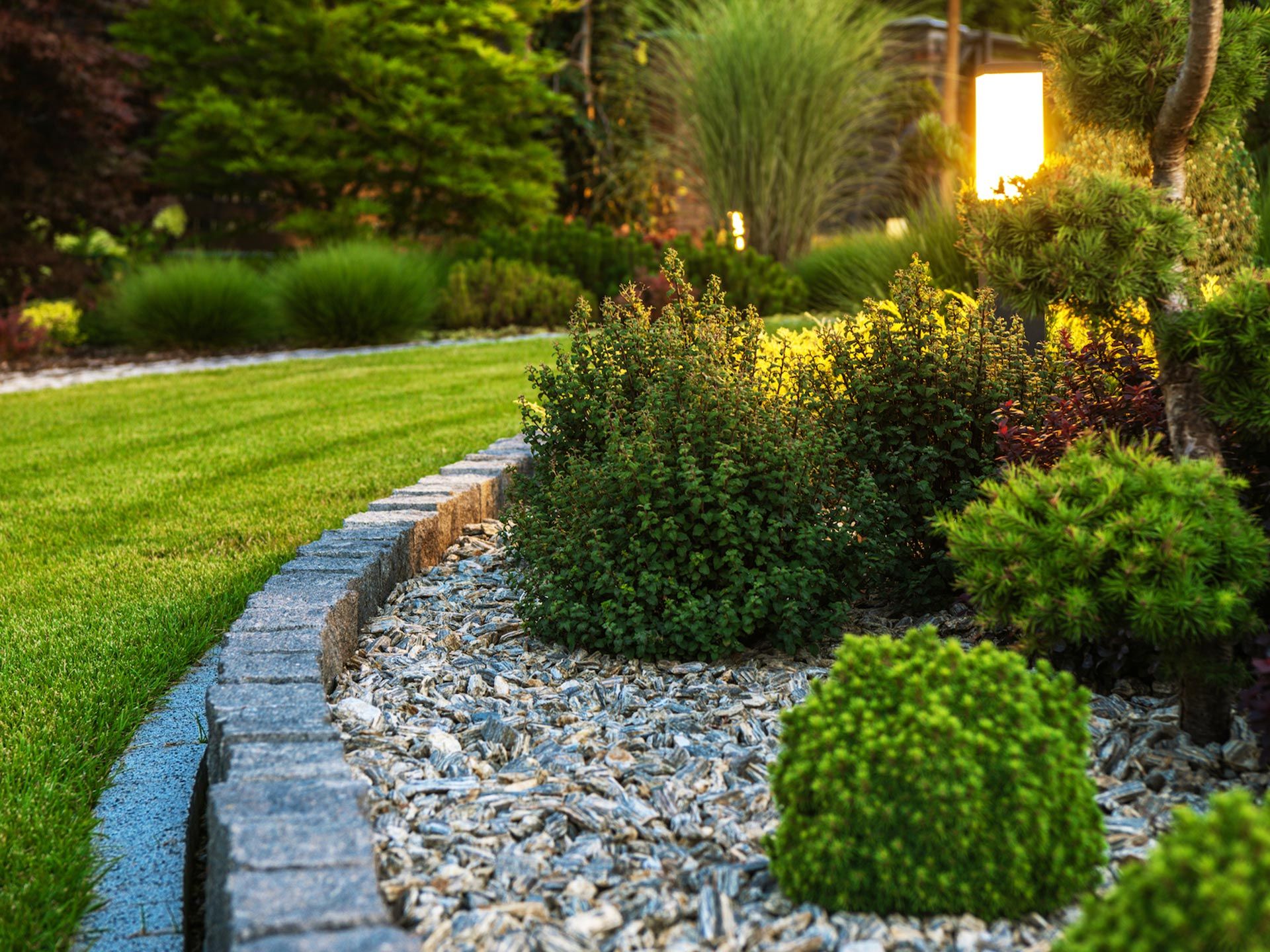 Lush garden bed with green shrubs and gravel, edged by a brick border and a lawn, illuminated by soft light.