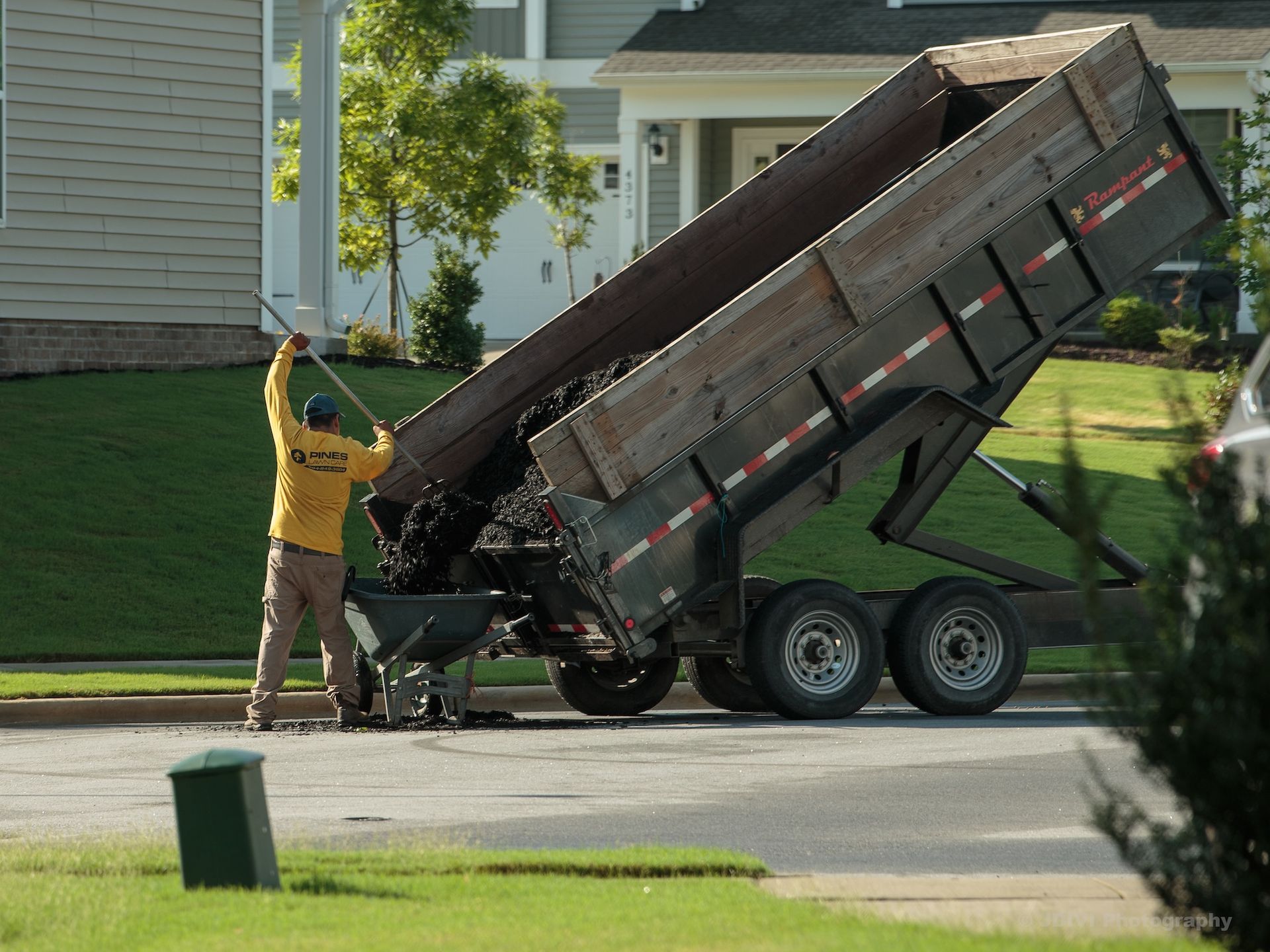 Man in yellow shirt unloading debris from a dump trailer onto a wheelbarrow on a residential street.