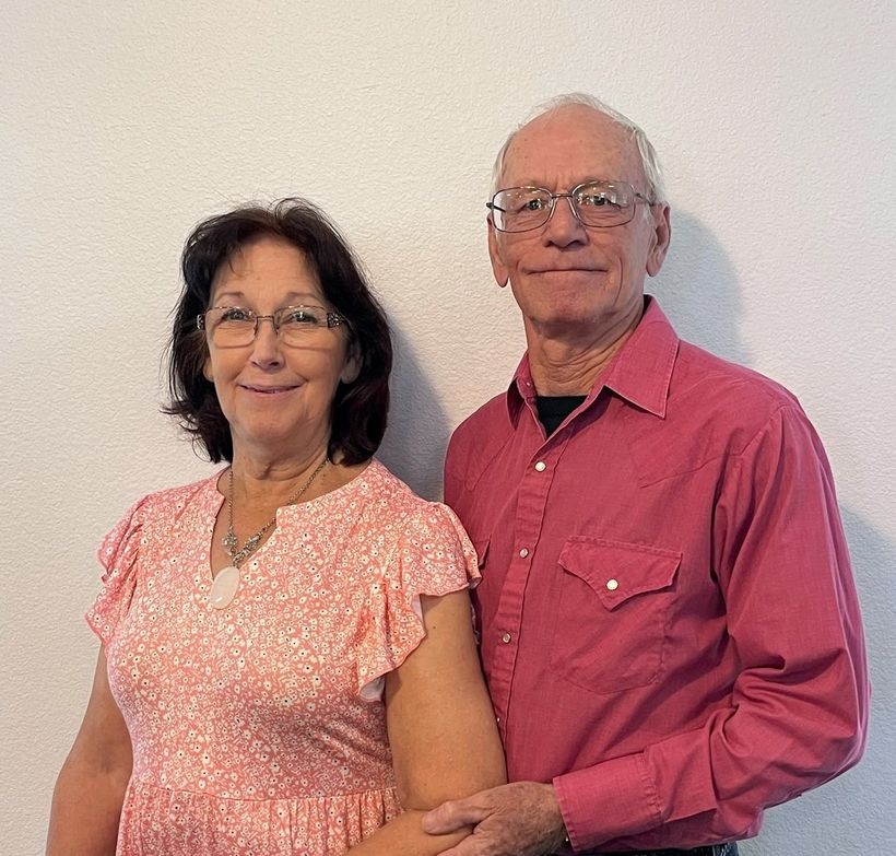 A smiling couple stands side-by-side against a plain white wall; the woman wears a pink floral dress and the man a red shirt.