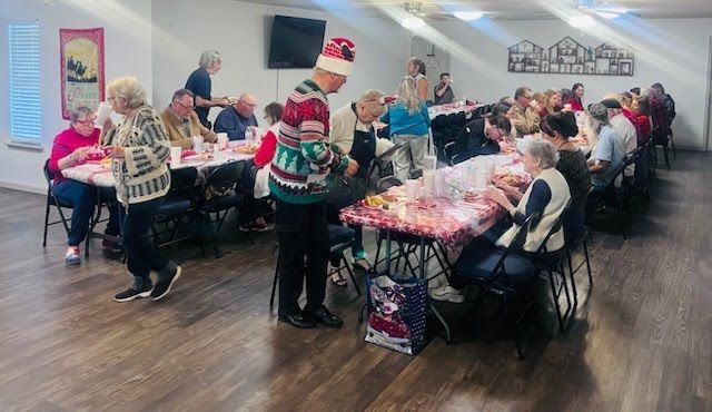 People at tables inside a community room, attending a holiday-themed gathering or festive event with shared food.