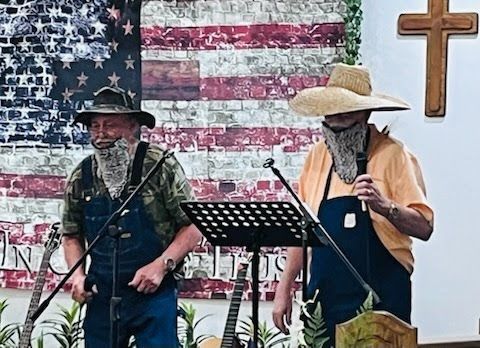Two individuals in overalls and beards perform with microphones in front of an American flag backdrop and a wooden cross.