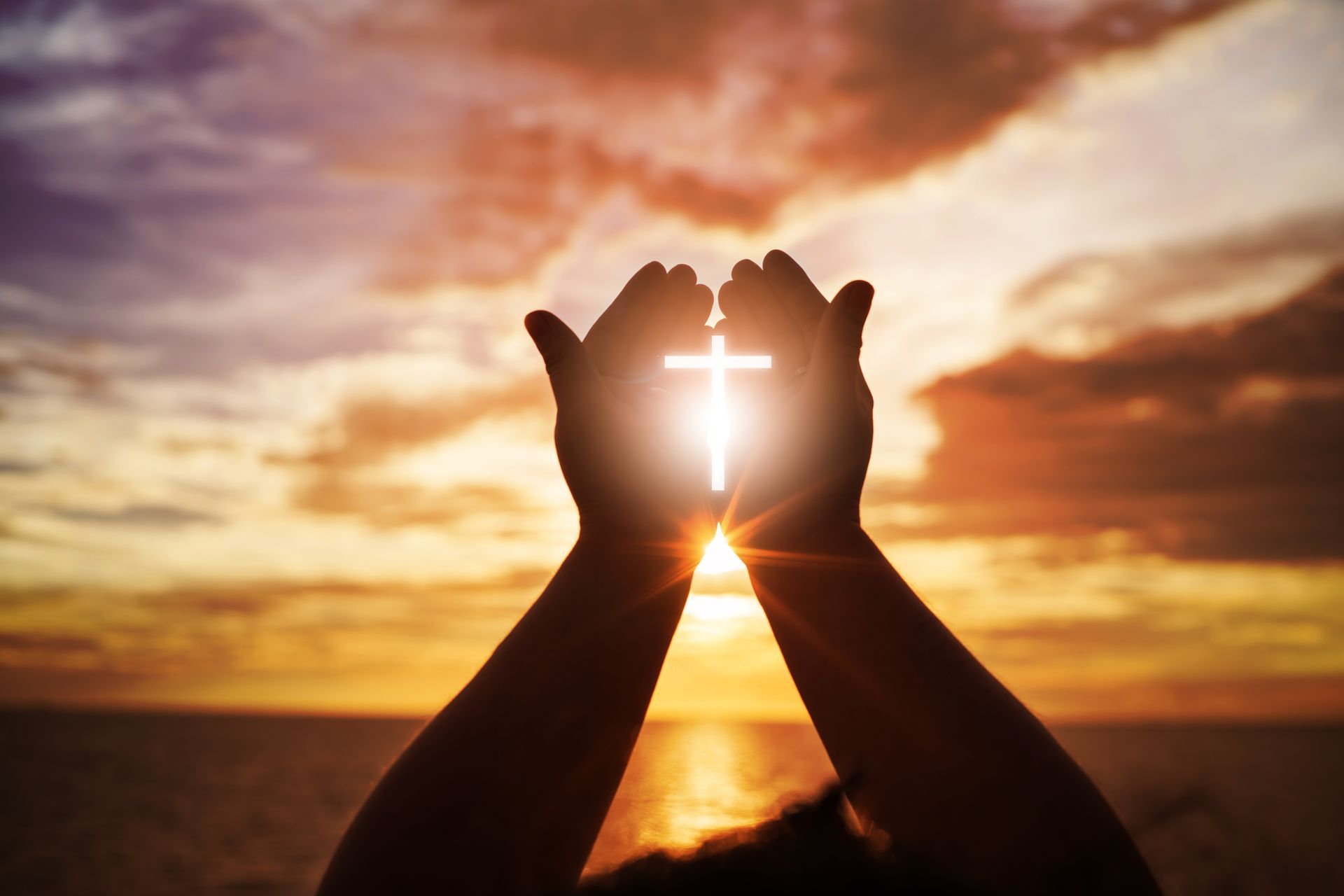 Silhouetted hands held upward against a sunset sky, forming a glowing, bright white cross between the palms.