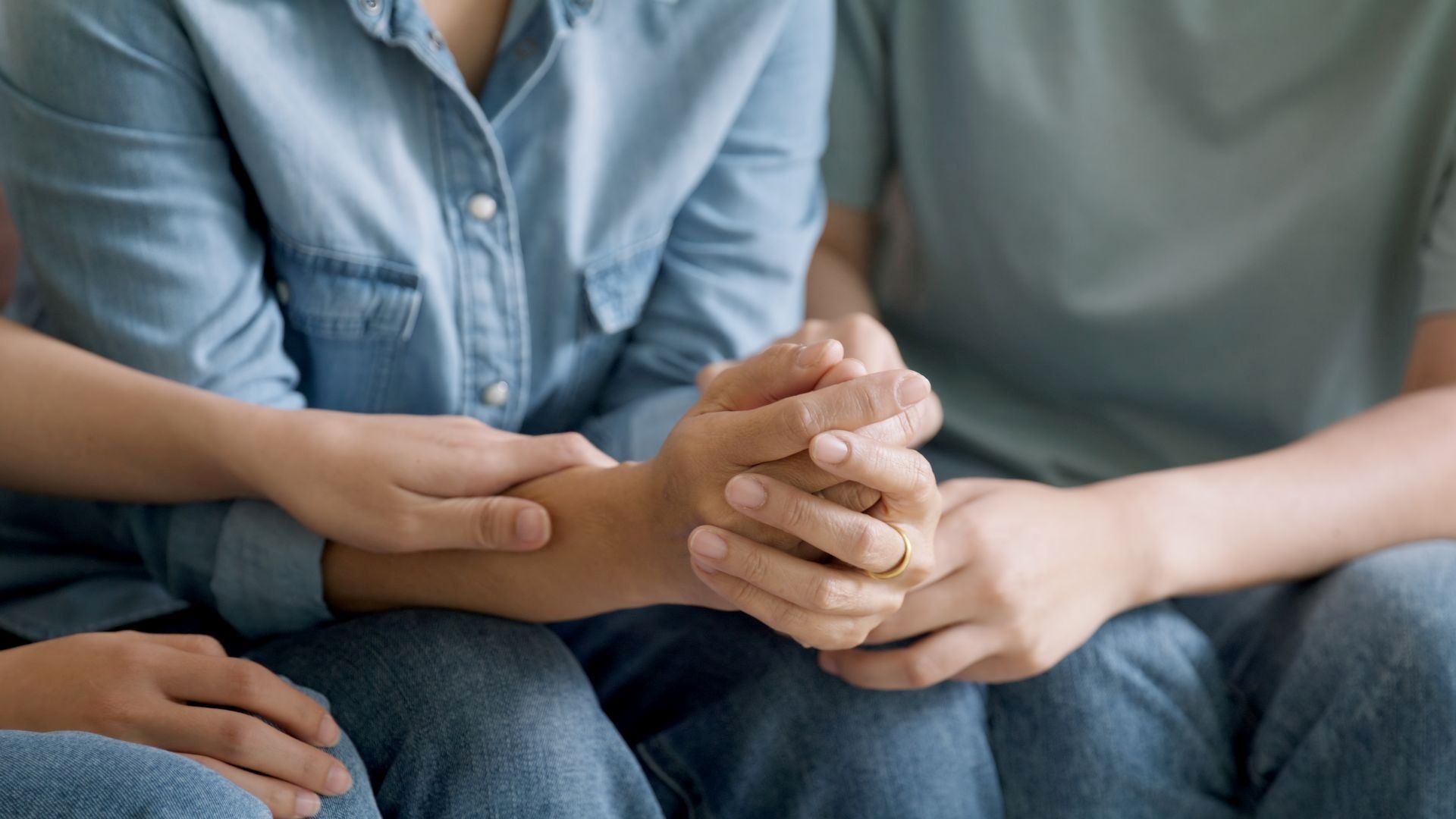 Two people hold hands, offering comfort. Close-up on arms, denim clothing, and a gold ring.