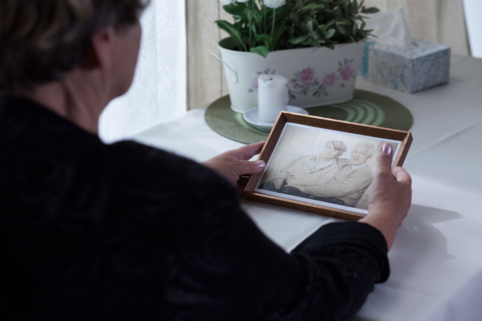 Person holding a framed photo, looking at it. White table, flowers, tissues, and a window are in the background.
