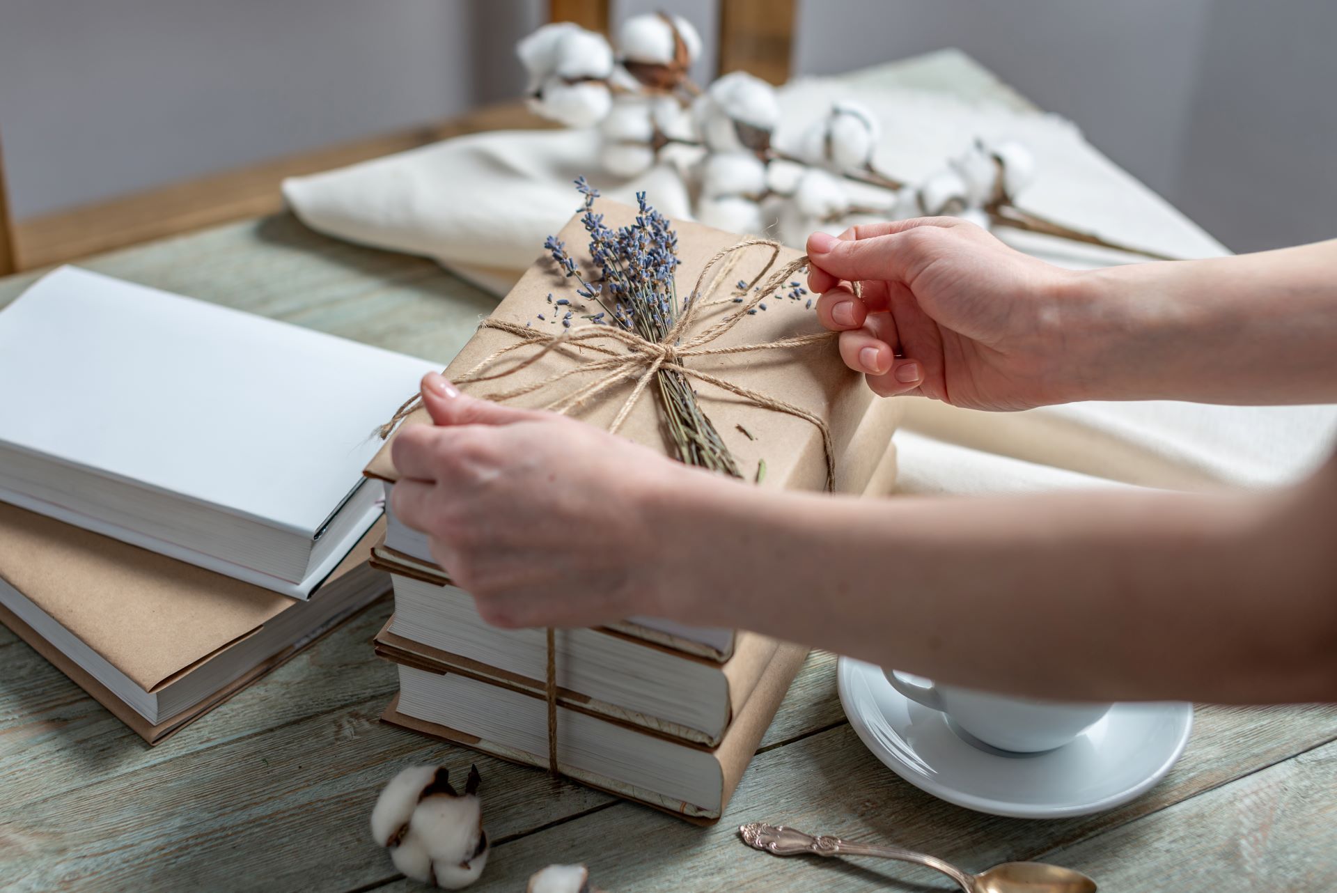 Hands tying twine around a stack of wrapped books, decorated with lavender and cotton, on a table.