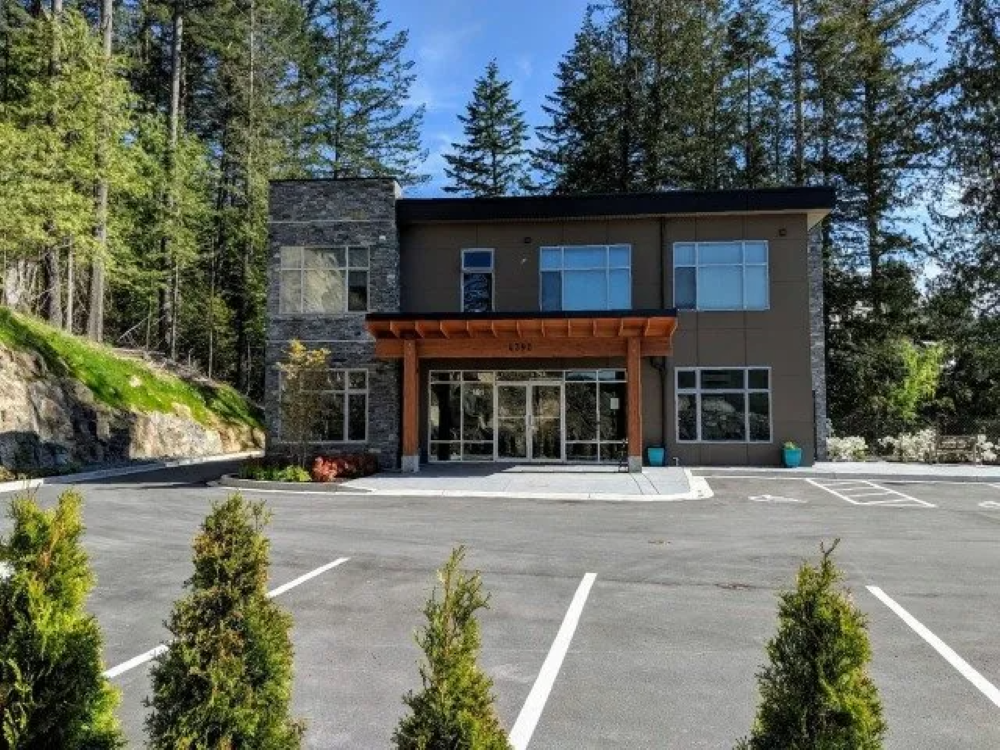 Modern building with stone accents, glass entrance, and wooden overhang. Surrounded by trees and parking lot.