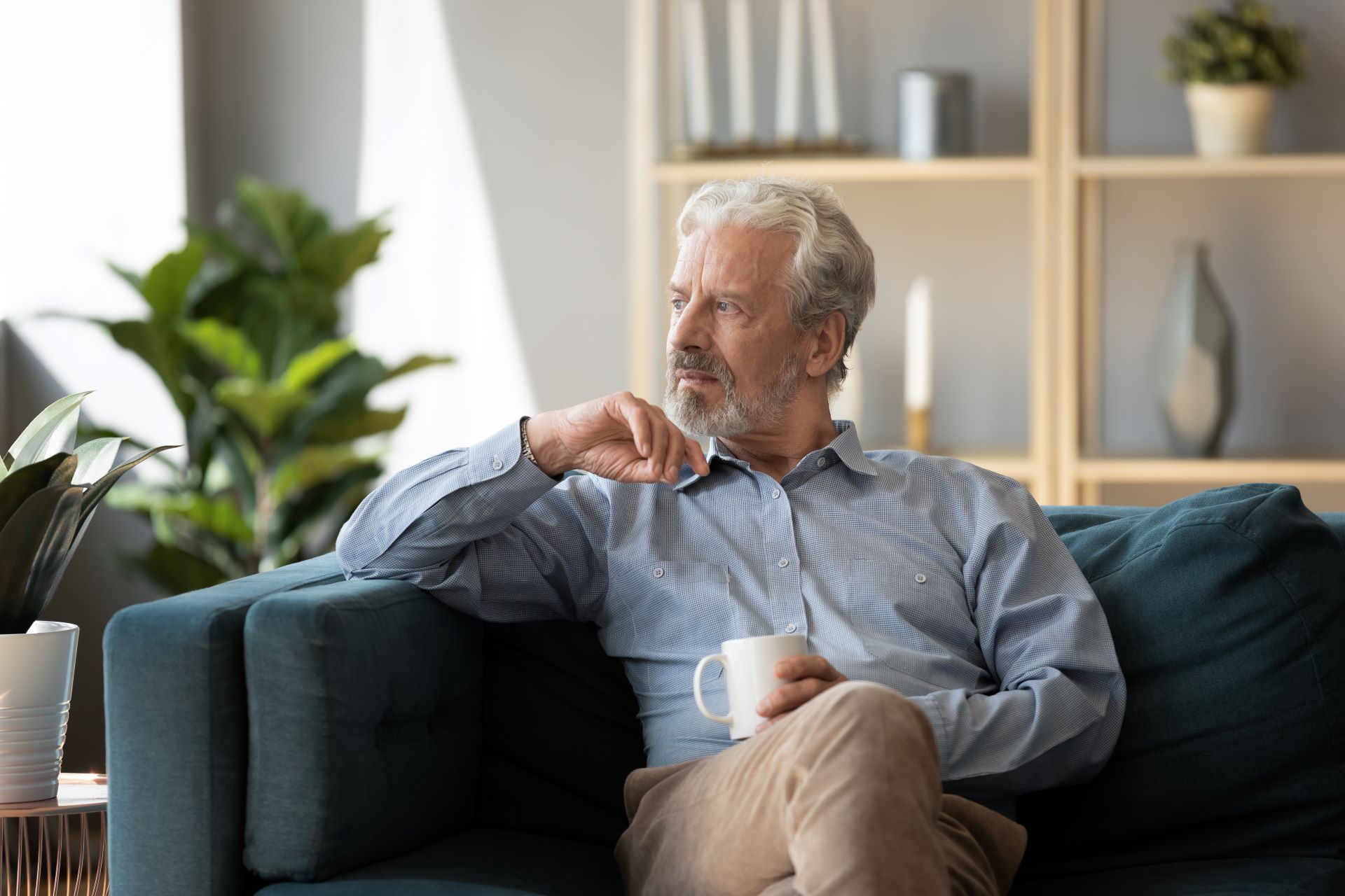 Older man with gray hair sits on a blue couch, holding a mug and gazing off. Interior setting with plants and shelves.