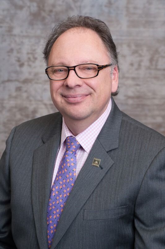 A man in a grey suit, light-colored striped shirt, and patterned purple tie, smiling against a mottled grey background.