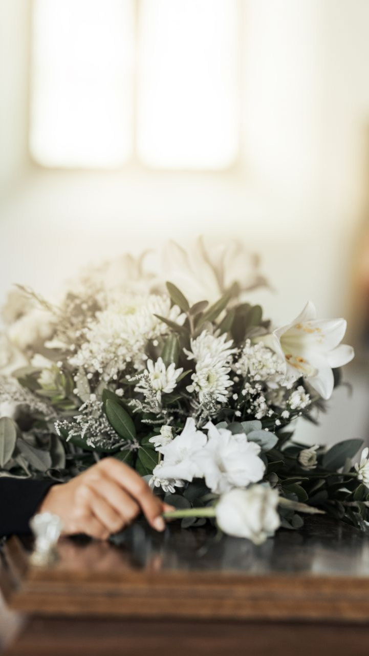 A hand rests on a wooden casket adorned with a large bouquet of white flowers, set against a soft, bright background.
