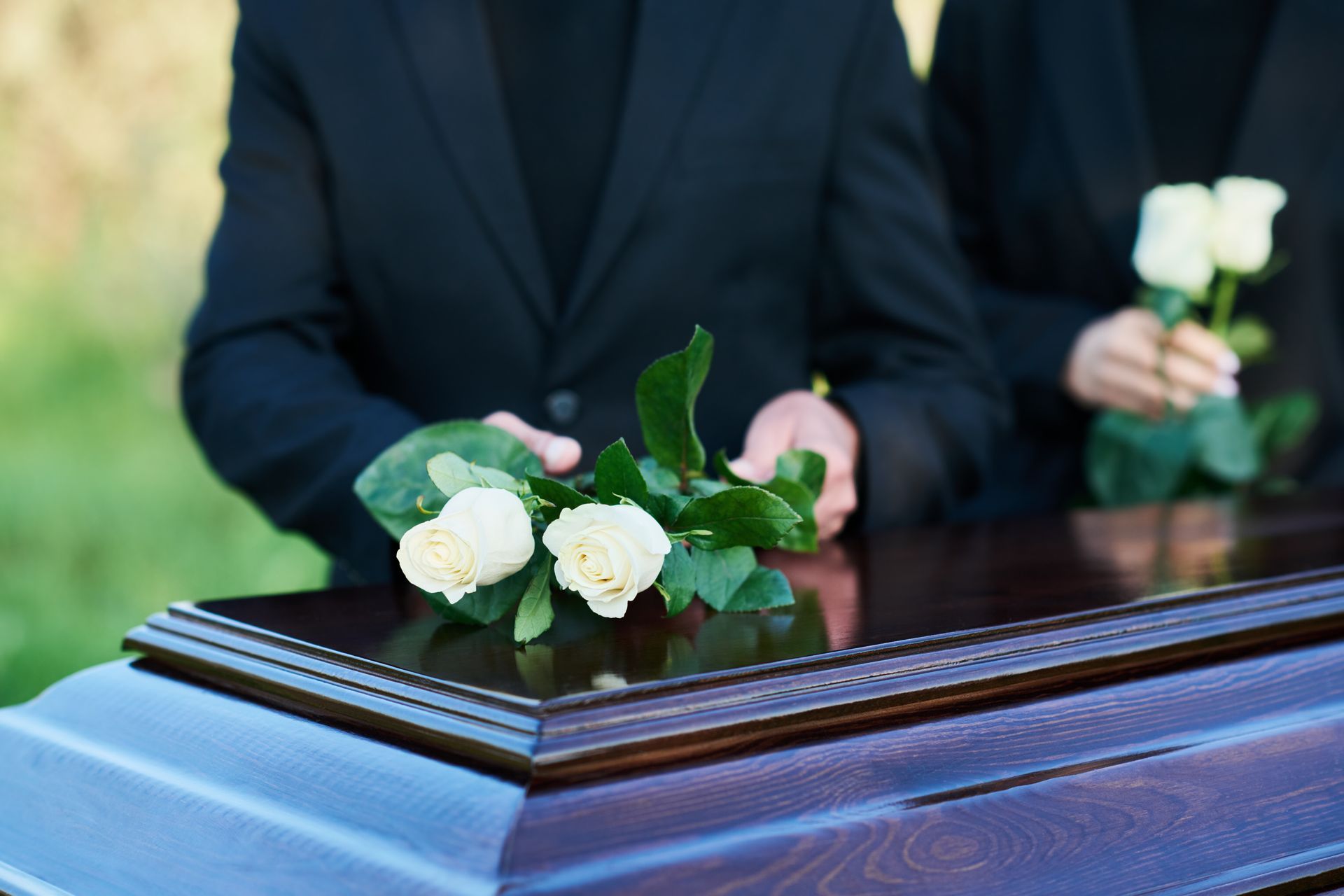 Two people in black suits place white roses on a dark wooden casket at an outdoor funeral service.