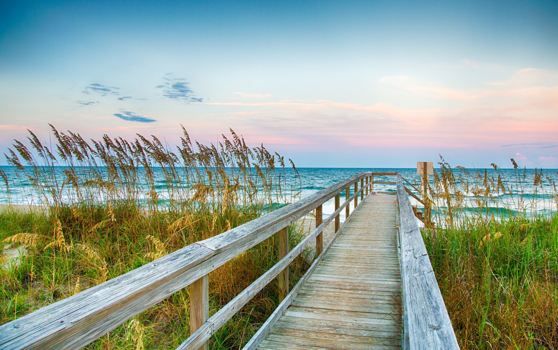A weathered wooden boardwalk leads through beach grasses toward the ocean under a pastel sunrise sky.