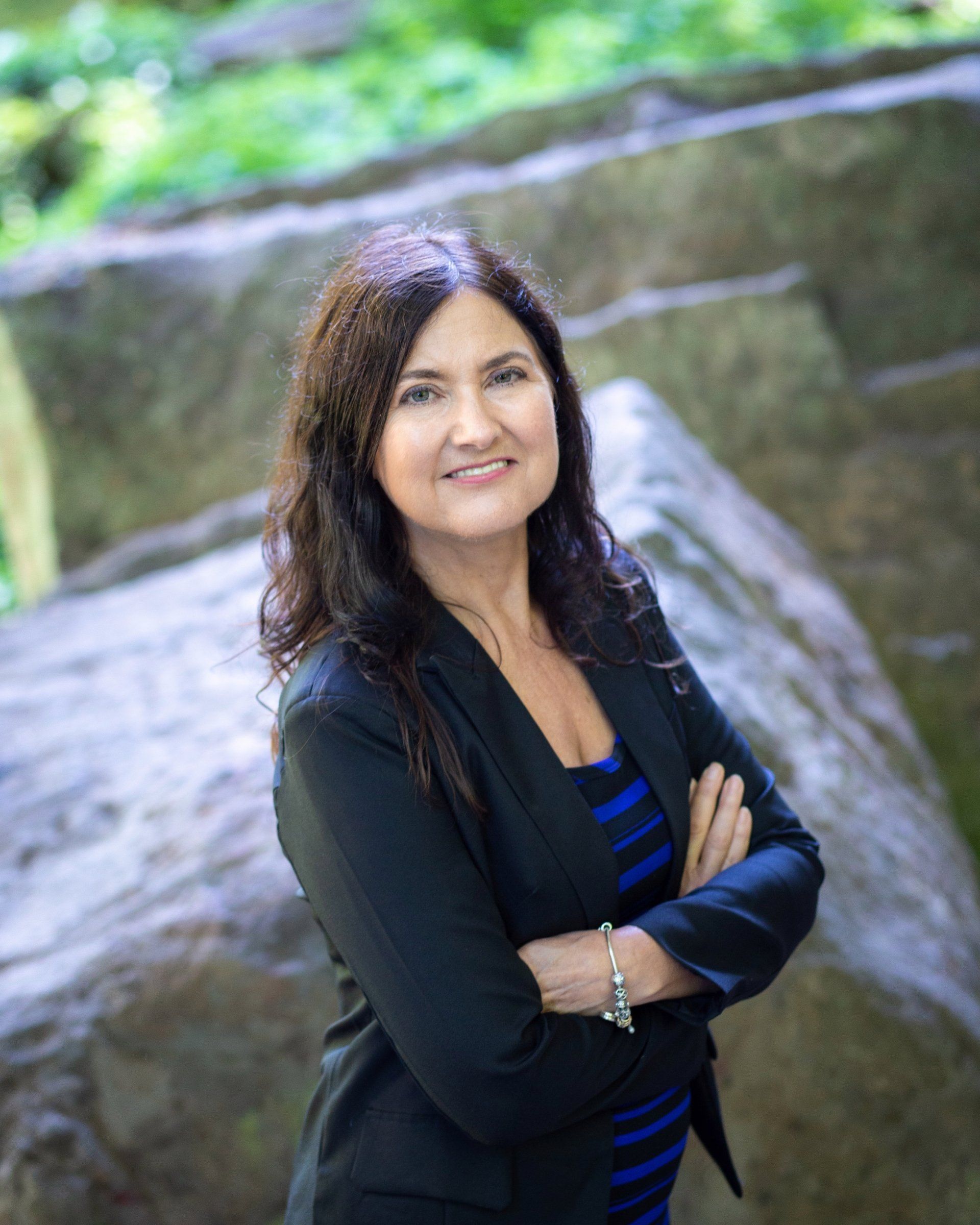 Woman with arms crossed, smiling, wearing a black blazer and blue striped top, standing in front of large rocks.
