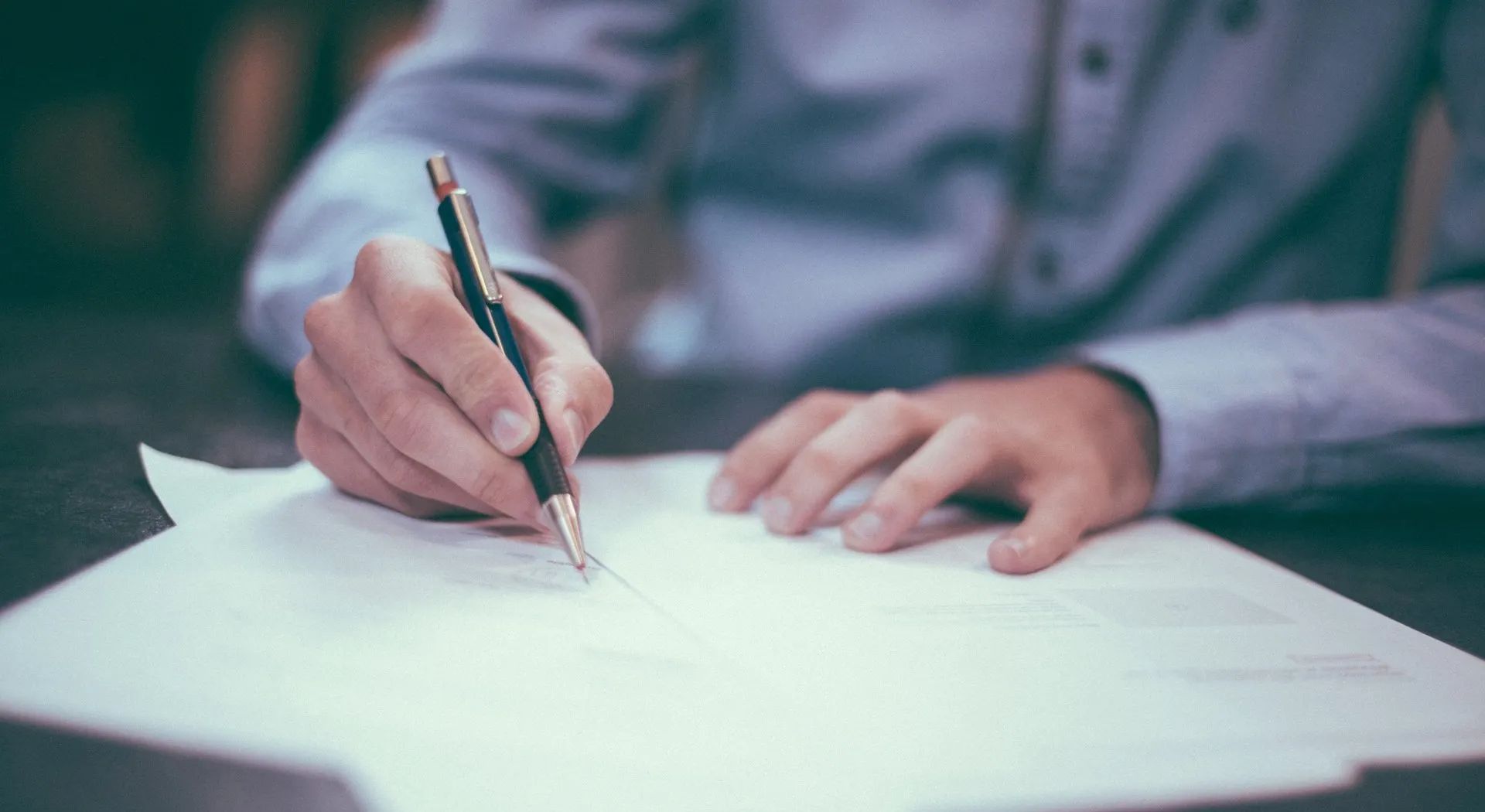 Person writing on a paper at a table; wearing a blue shirt and holding a pen.