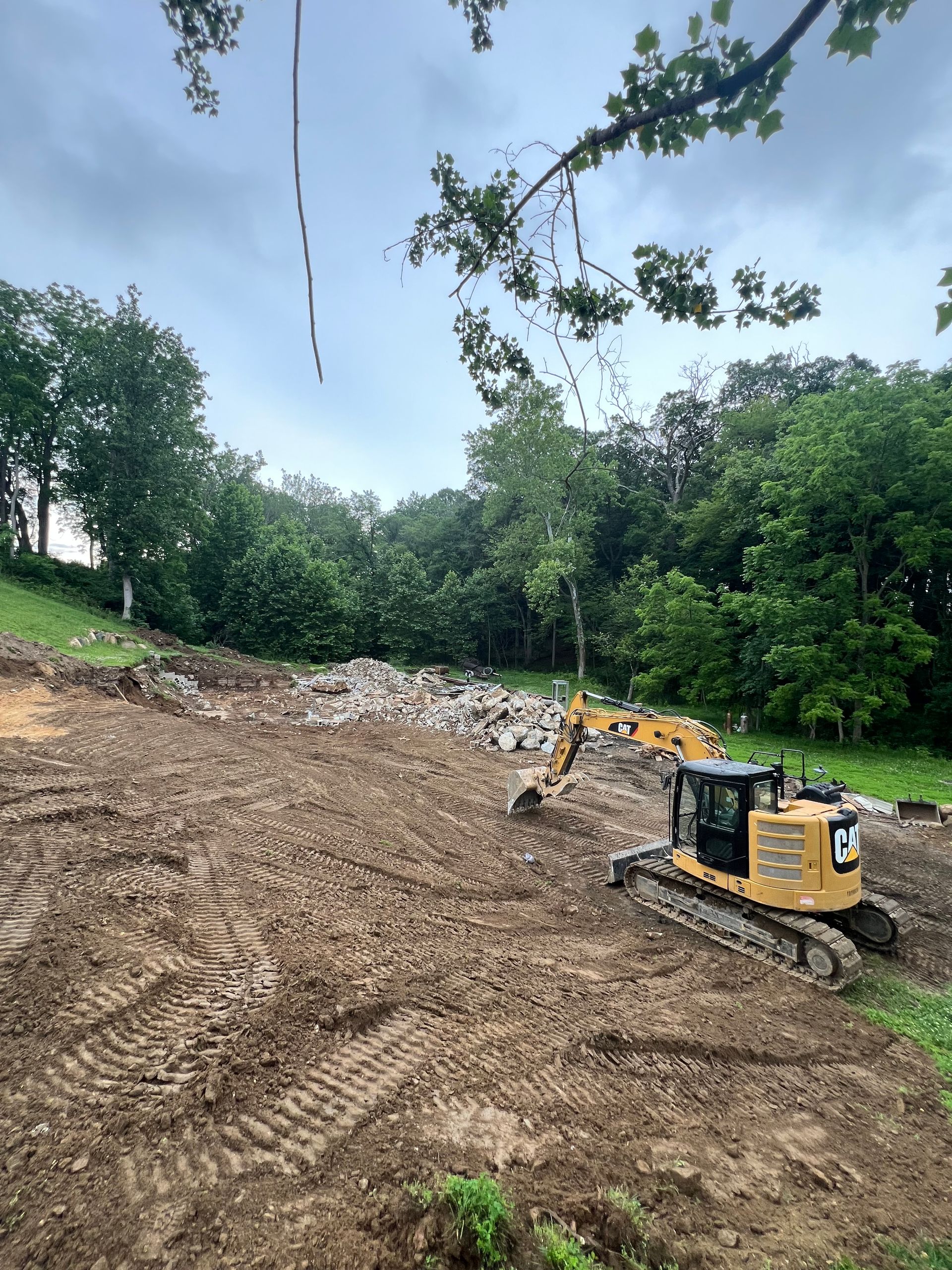 Excavator clearing a muddy hillside with piles of debris under a cloudy sky, surrounded by trees