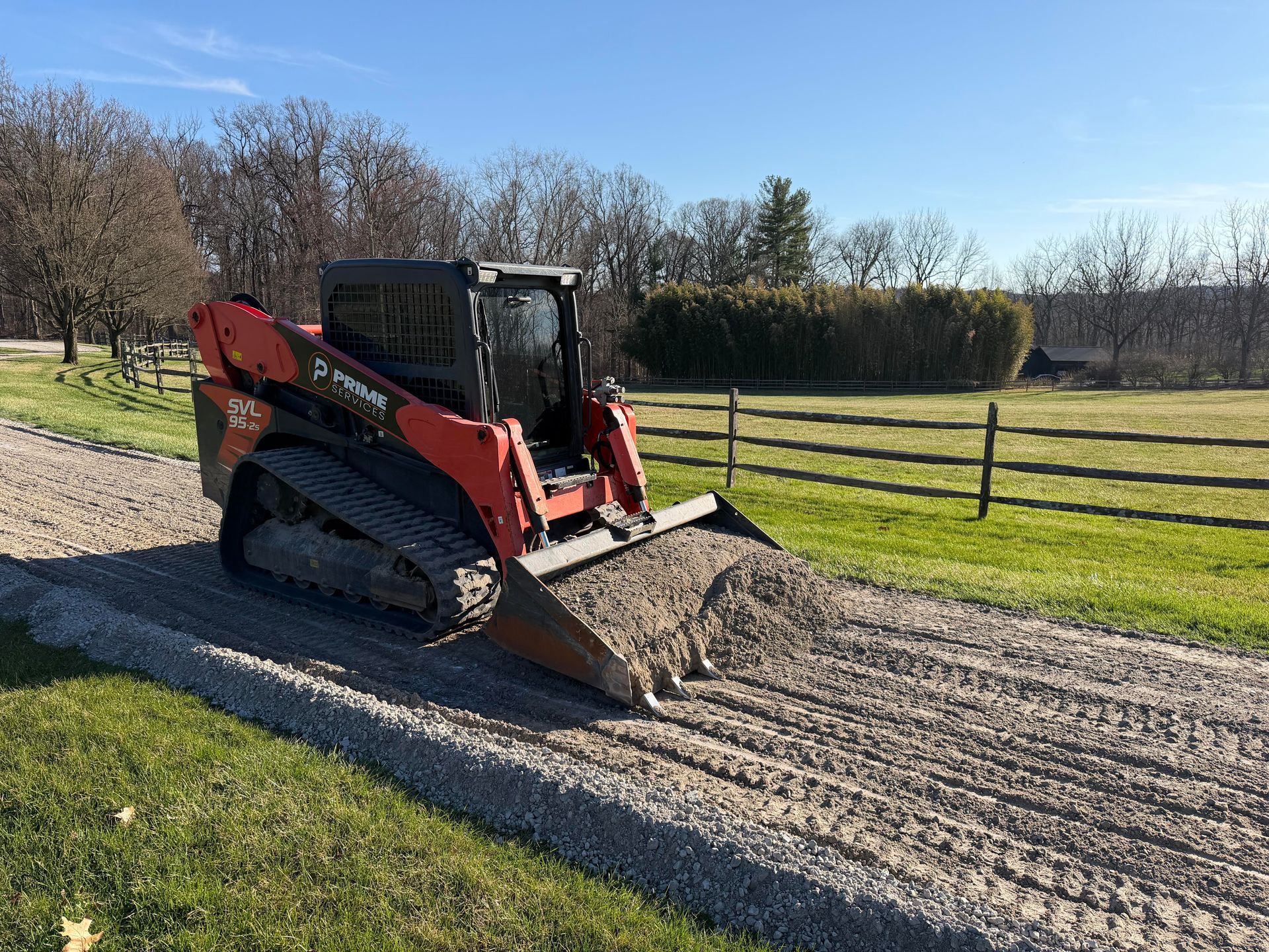 Red compact tractor with front loader grading a gravel driveway beside a grassy field and fence.